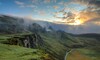 Weitläufige, grün bewachsene bergige Landschaft, über der Sonnenstrahlen aus einer Wolkendecke hervorkommen